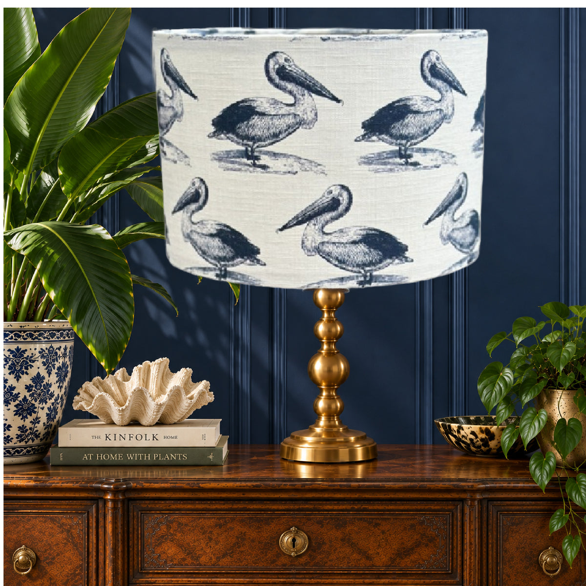 A table lamp with a white drum shade printed with blue pelican illustrations and a polished gold-toned metal base, styled on a wooden chest against a dark blue paneled wall with plants and decor.