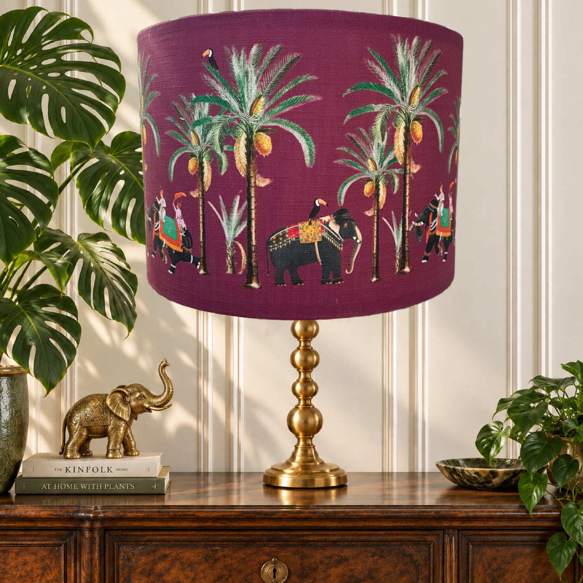 A table lamp with a deep purple fabric drum shade featuring a detailed print of palm trees, elephants, and riders, supported by a polished gold brass base. The lamp is styled on a dark wood cabinet next to a potted monstera plant and a small gold elephant figurine.