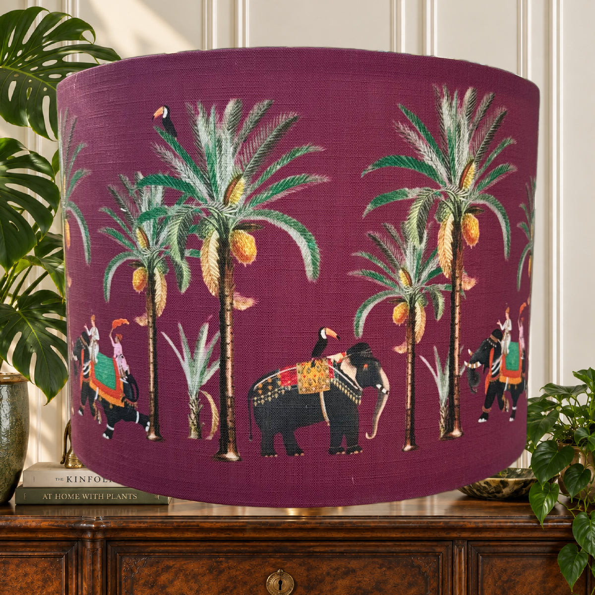 A fabric drum lampshade with a deep magenta-purple background, decorated with a colorful tropical print featuring palm trees, toucans, and elephants. The lampshade is styled on a wooden cabinet with house plants in the background.